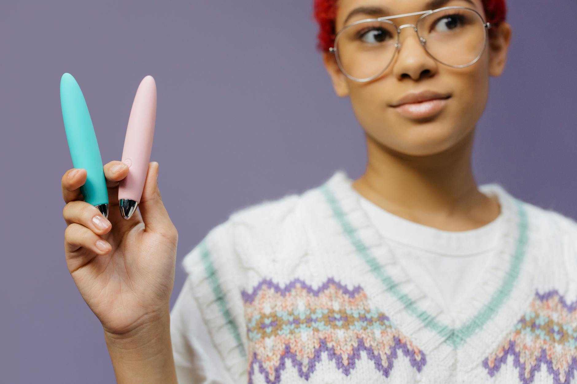 A person with eyeglasses holding two colorful vibrators against a purple background